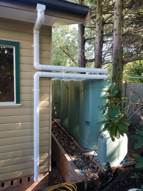 New PVC downpipe running from the gutter of a weatherboard house, with charged lines carried across the tops of a bank of green poly rainwater tanks