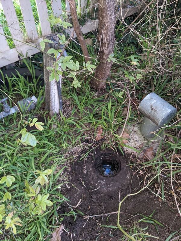 The same opening with nothing in it and no cover in place, standing water in the bottom, sitting in a saucer of bare dug soil with grass at its outer edge