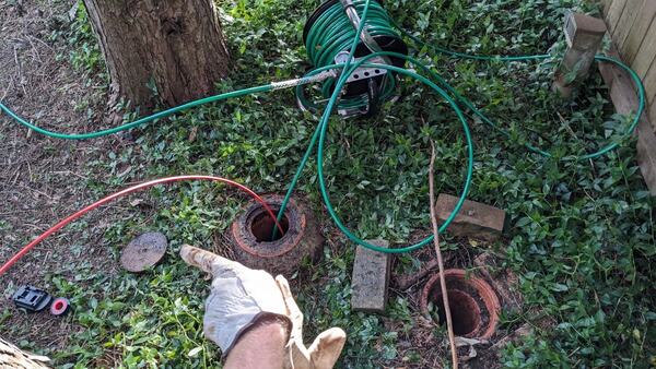 Two open earthenware drain inspection openings set in low ground cover, with a green hose and a thin red line fed into one of them, a gloved hand in the foreground, a stainless hose reel behind and a concrete lid lifted aside