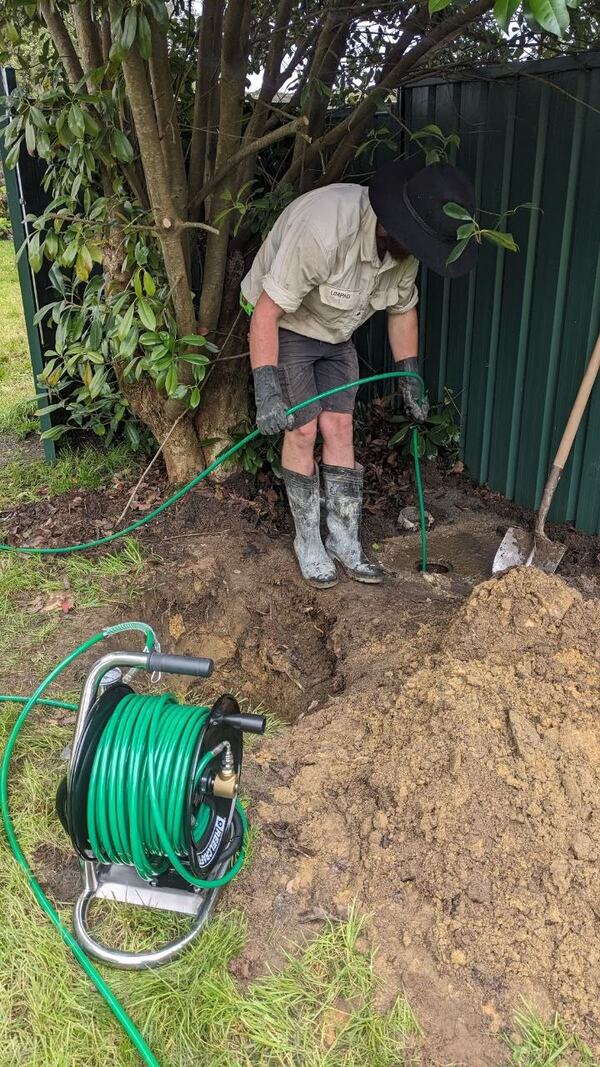 A plumber in an Upper Mountains Plumbing and Gas shirt and wide-brimmed hat bending to feed a green hose into a small opening in the ground beside a colourbond fence, with a stainless hose reel, a heap of excavated soil and a shovel beside him
