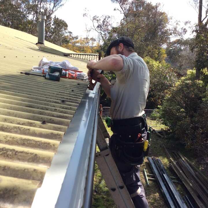 Plumber on a ladder fitting a new length of gutter along the edge of a corrugated metal roof