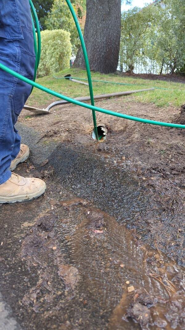 A green hose fed into the broken end of a pipe in a shallow excavation, with muddy water and silt running out of the pipe and across the ground