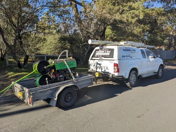 A green high-pressure drain jetter with twin hose reels on a box trailer behind a signwritten Upper Mountains Plumbing and Gas ute, stopped on a quiet bitumen street with the jetting hose run off the trailer and away over the grass verge