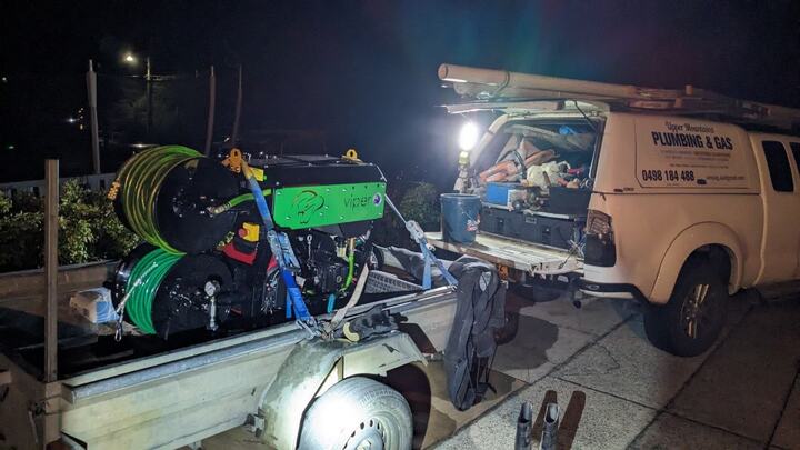 A green high-pressure drain jetter with twin hose reels strapped to a box trailer at night, lit by a work light, alongside the Upper Mountains Plumbing and Gas ute with its tailgate down, the canopy open and tools inside