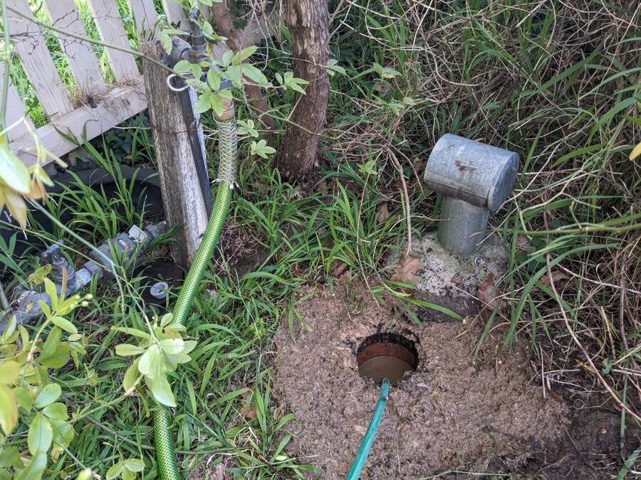A green hose fed into a round opening in bare soil in an overgrown garden bed, with a galvanised pipe set in a concrete pad to one side and a tap on a galvanised riser beside a timber fence post behind