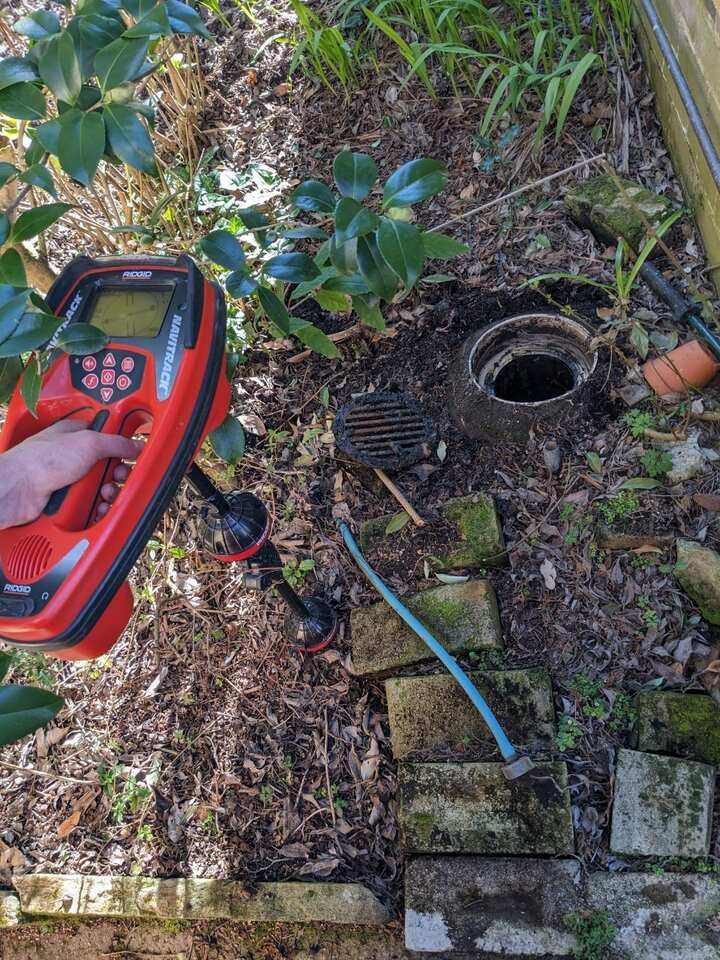 Plumber holding a RIDGID NaviTrack pipe locator beside an opened inspection point in a garden bed