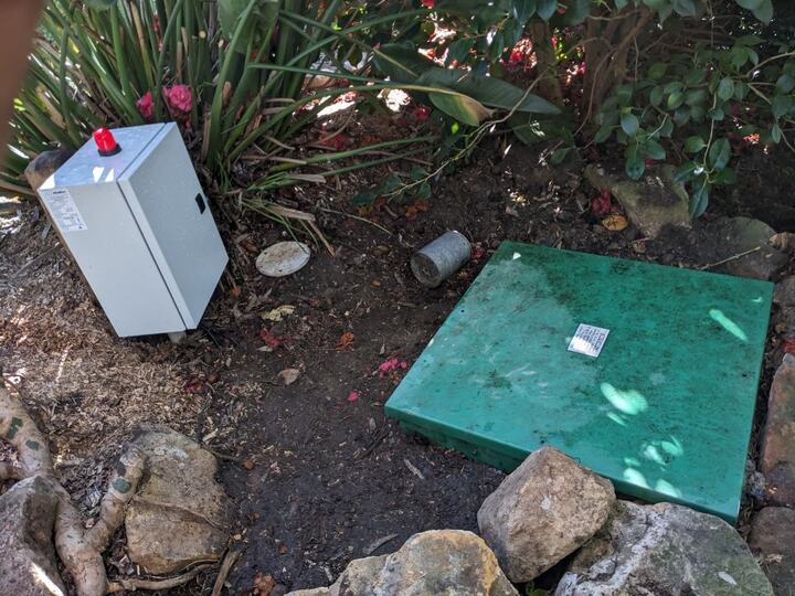 Angled view of a finished pump-out installation in a garden bed, showing the white control cabinet with its red alarm beacon beside the closed square green well lid