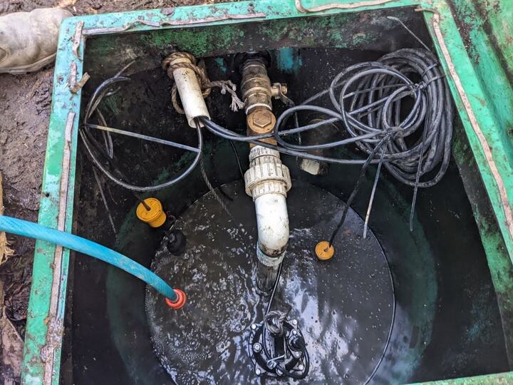 Looking down into an open wet well at a submersible sewage pump sitting in effluent on a lifting rope, with its rising main coming up through a barrel union, two orange float switches on their cables and a blue hose run into the well