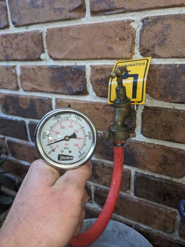 Pressure gauge hosed onto a brass garden tap, beneath the yellow RAINWATER tag that marks a rainwater-fed outlet