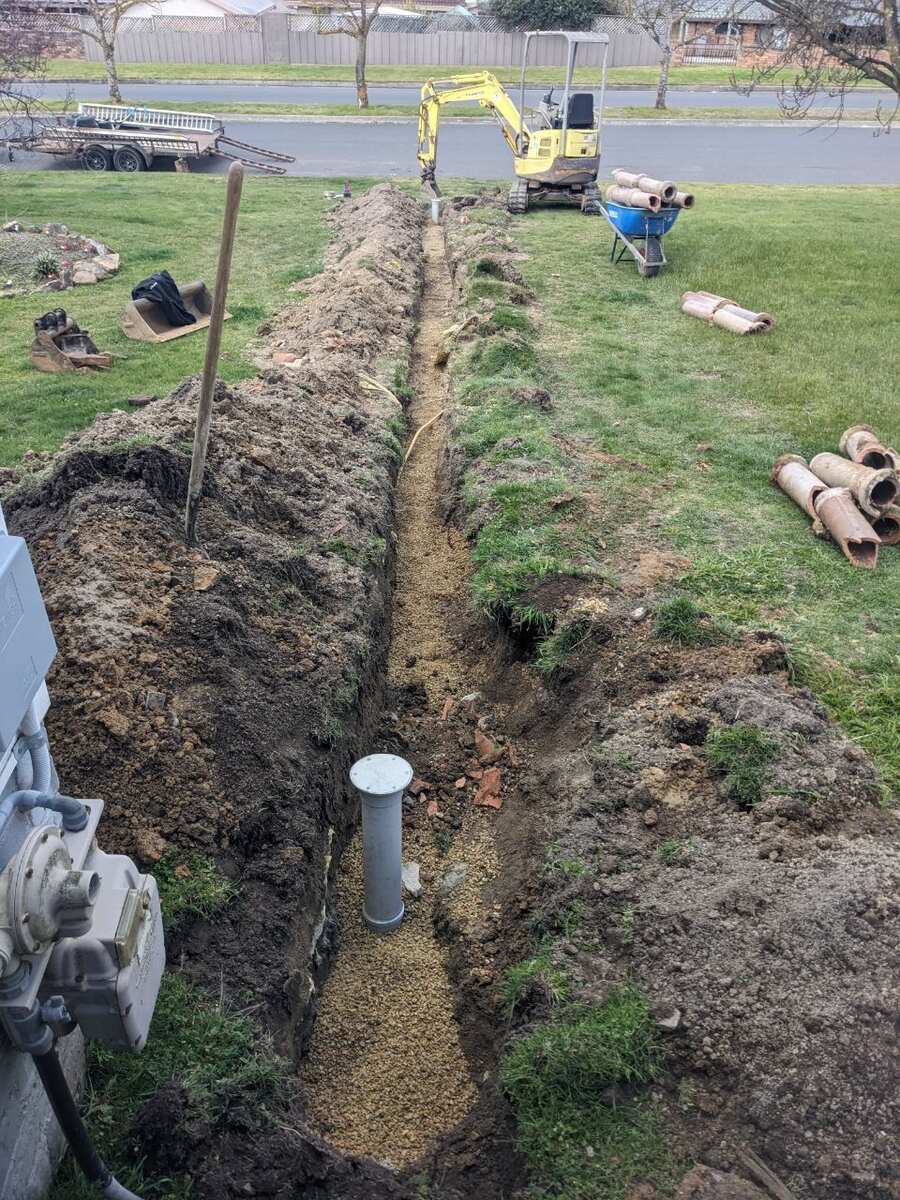 Long open trench across a front lawn with a mini excavator at the far end, lengths of old earthenware pipe stacked on the grass, and a new inspection riser set in gravel