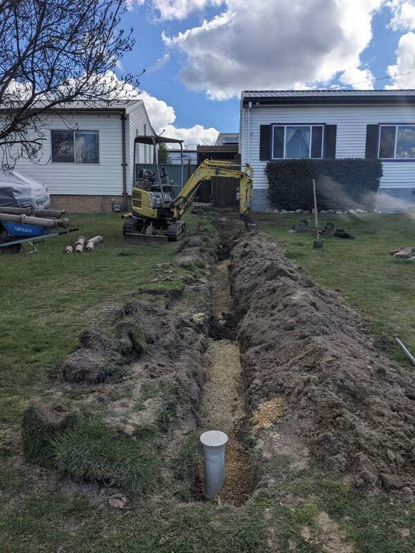 Open trench running between two houses with a mini excavator at the far end and a new inspection riser standing in the gravel bed