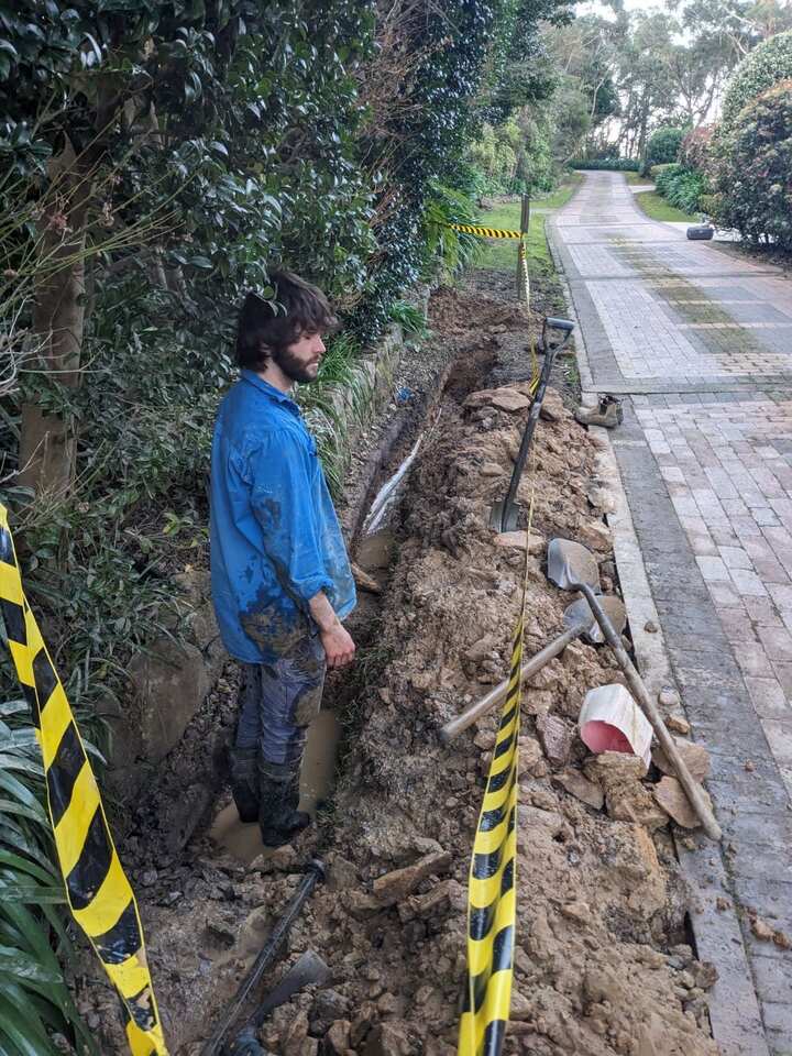 Plumber standing in a narrow trench dug alongside a driveway, with barrier tape either side and a new water line in the bottom of the trench